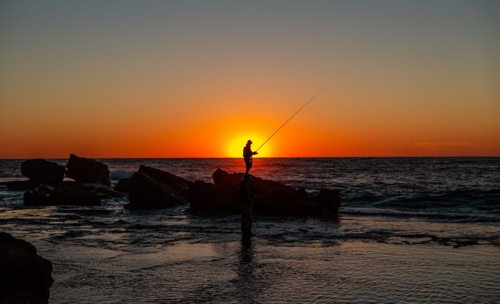 Una persona de pie sobre una roca junto a la orilla, pescando mientras el sol se pone a sus espaldas